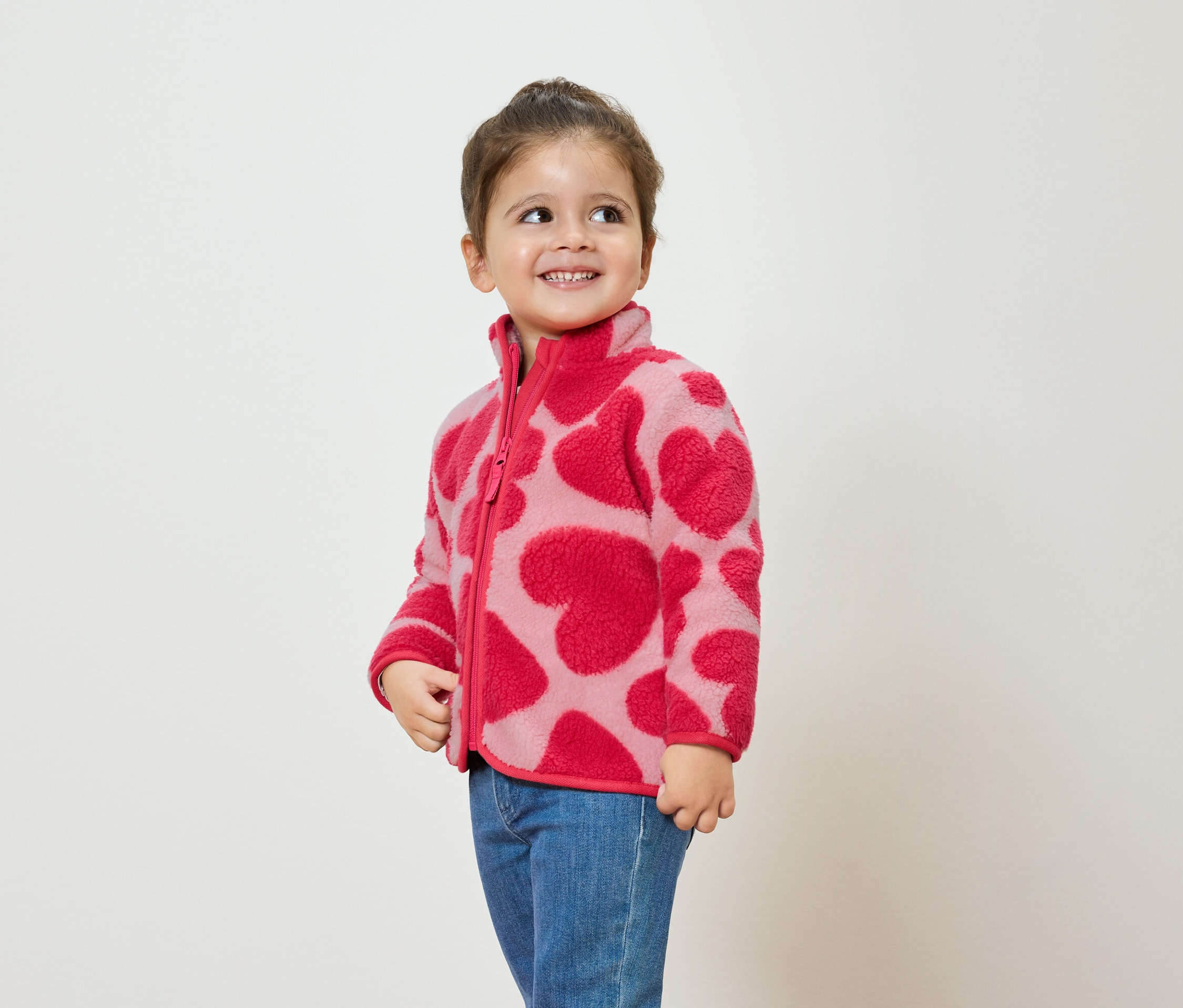 Fillette portant une veste en polaire rose à motifs de cœurs et un jean bleu, souriant et regardant sur le côté.