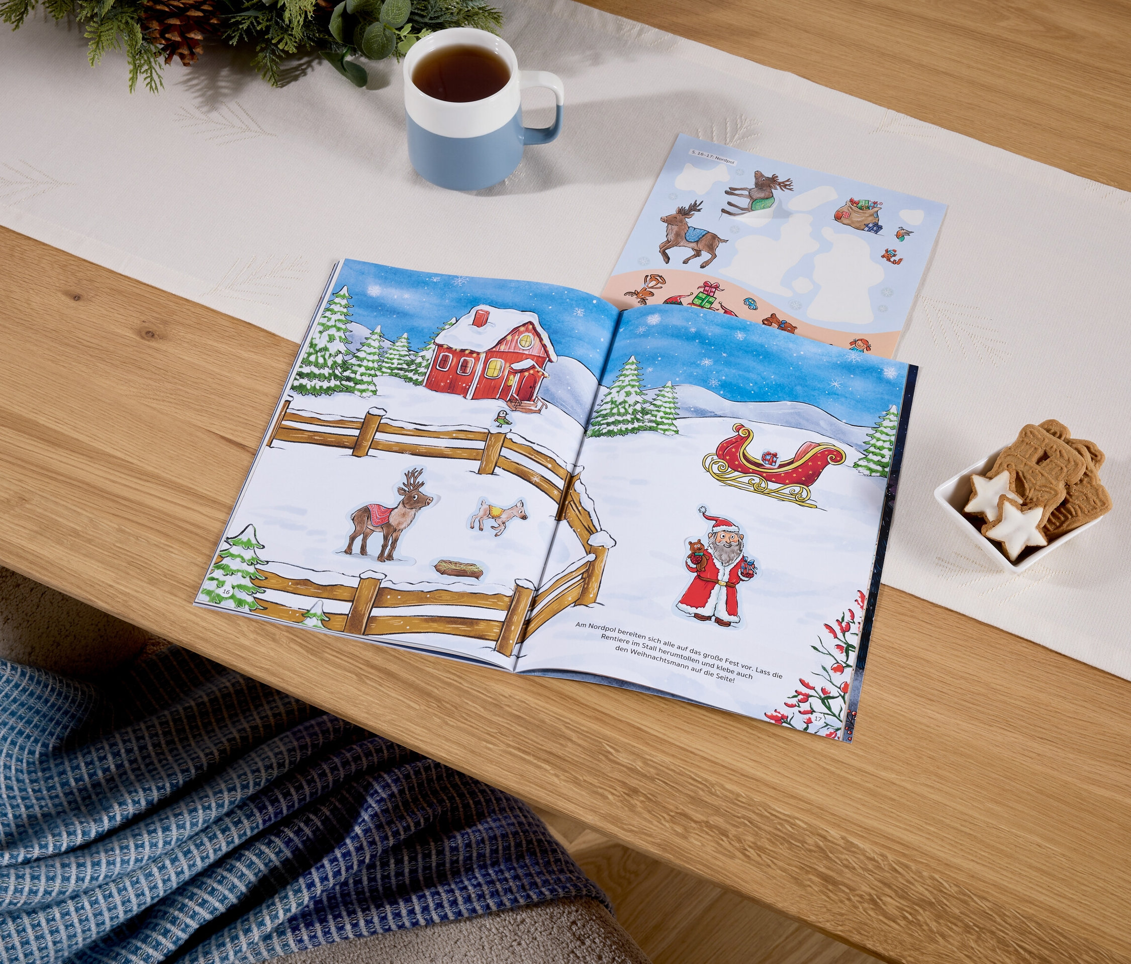 Sur une table, un livre ouvert illustré sur le thème de Noël, à côté d'une tasse avec une boisson et d'un bol de biscuits.
