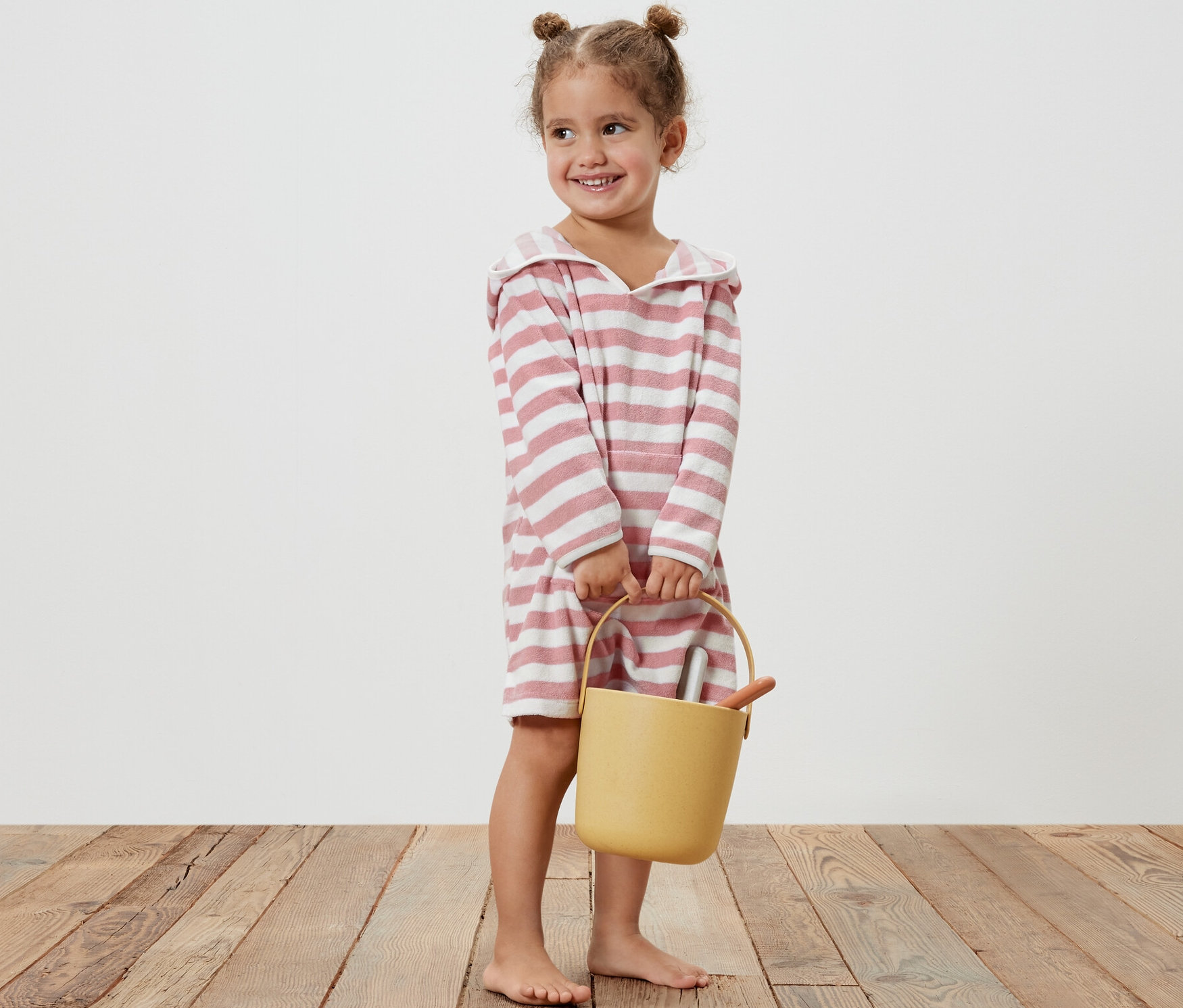 Petite fille souriante tenant un seau jaune et portant un poncho en tissu éponge pour enfant à rayures roses.