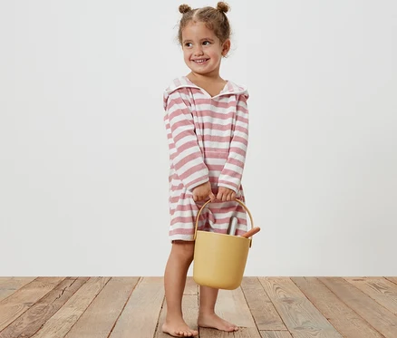 Petite fille souriante tenant un seau jaune et portant un poncho en tissu éponge pour enfant à rayures roses.