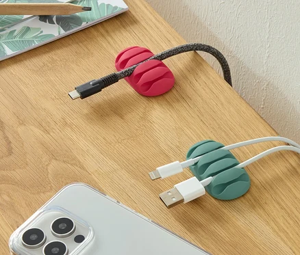 Sur une table, il y a deux range-câbles universels, un rose et un vert, ainsi qu'un téléphone et des crayons.