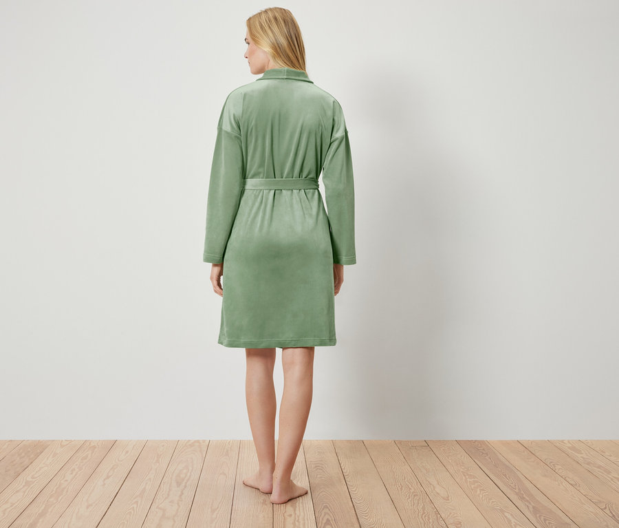 Vue de dos d'une femme blonde portant un peignoir vert clair, debout sur un plancher en bois devant un mur blanc.