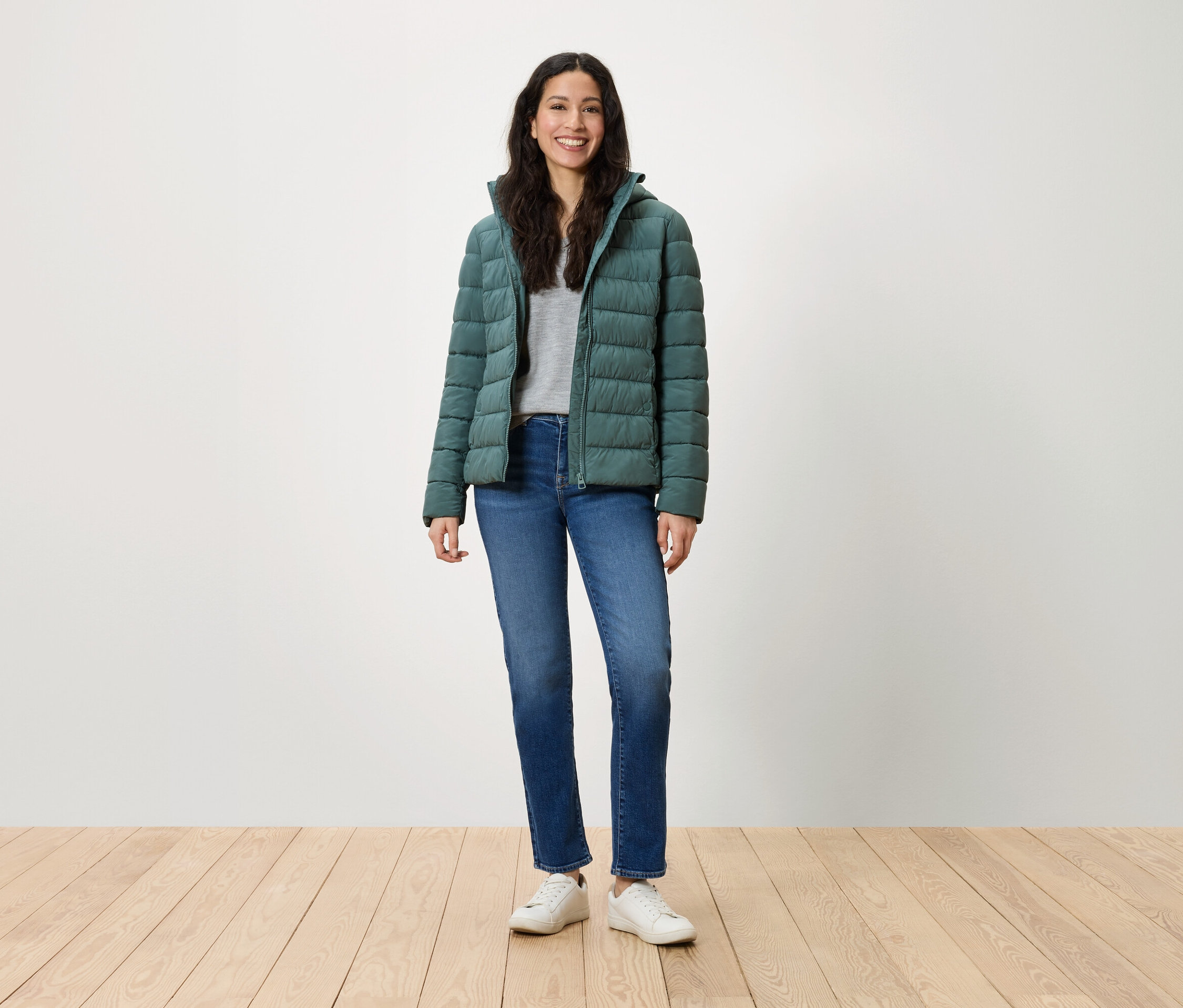 Une femme se tient debout et sourit, portant une doudoune verte, un jean bleu et des baskets blanches.