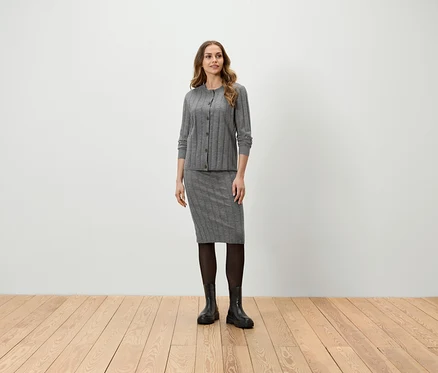 Une femme debout portant un cardigan gris, une jupe, des collants noirs et des bottes.
