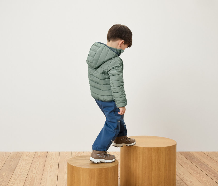 Un garçon en veste verte et pantalon bleu monte sur un socle en bois.