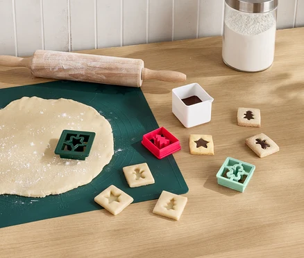 Un tapis vert avec de la pâte étalée, un rouleau à pâtisserie et un emporte-pièce en forme d'étoile. À côté se trouvent des biscuits découpés avec du chocolat et des emporte-pièces en forme d'arbre, d'étoile et de bonhomme.