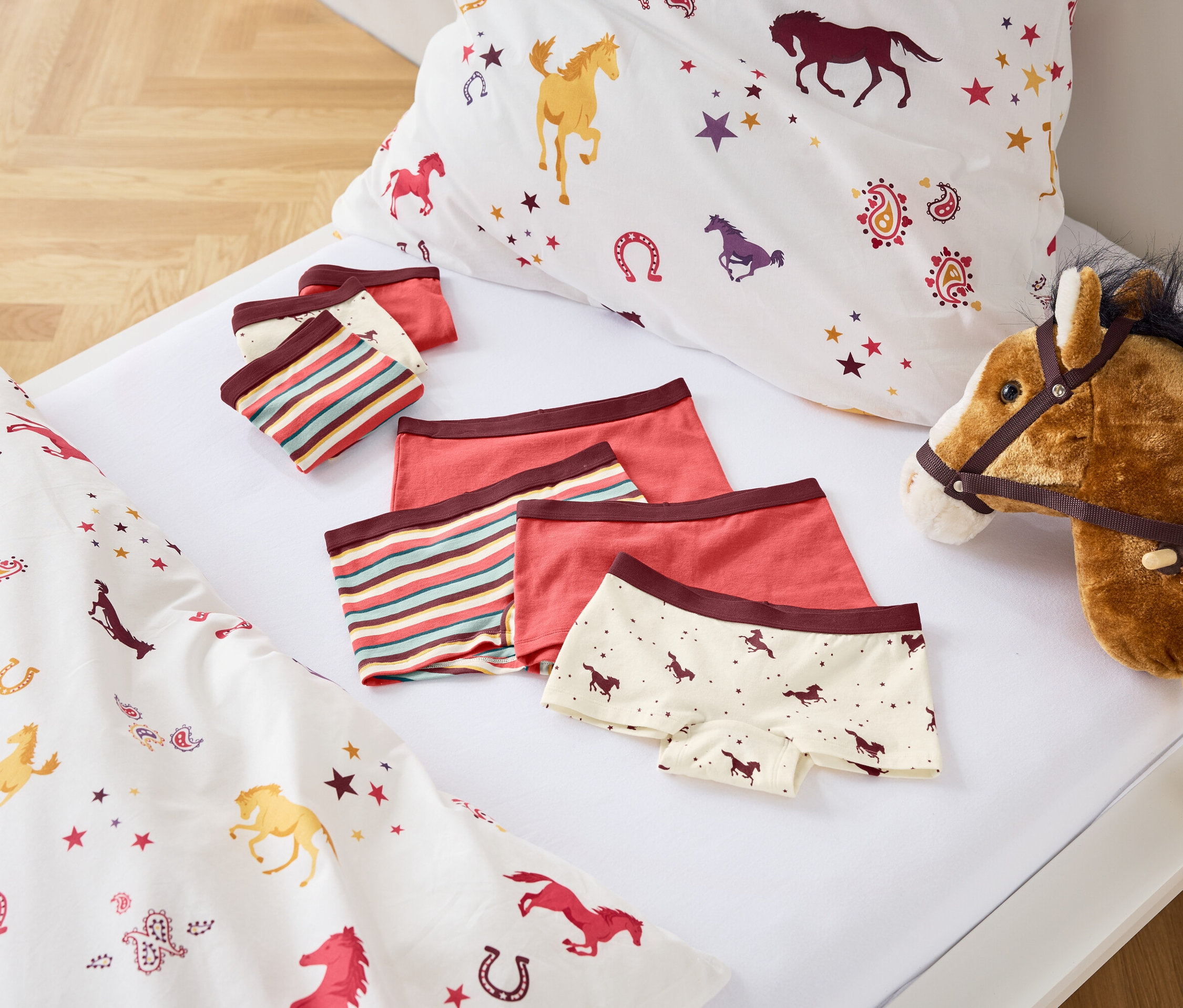 Sur un lit blanc sont disposés des draps imprimés de motifs de chevaux, un ensemble de sous-vêtements pour enfants et un cheval sur bâton.