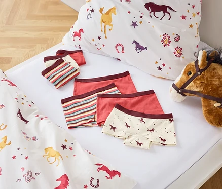 Sur un lit blanc sont disposés des draps imprimés de motifs de chevaux, un ensemble de sous-vêtements pour enfants et un cheval sur bâton.
