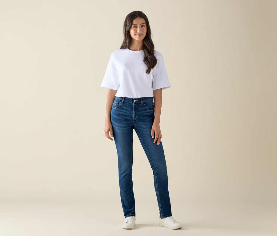 Une femme debout porte un jean slim – Coupe « Emma », denim bleu moyen et un t-shirt blanc.