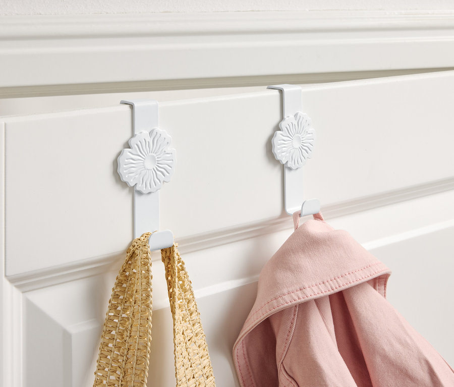 Deux crochets blancs avec un motif floral suspendus à une porte blanche, sur lesquels sont accrochés un sac jaune et une veste rose.
