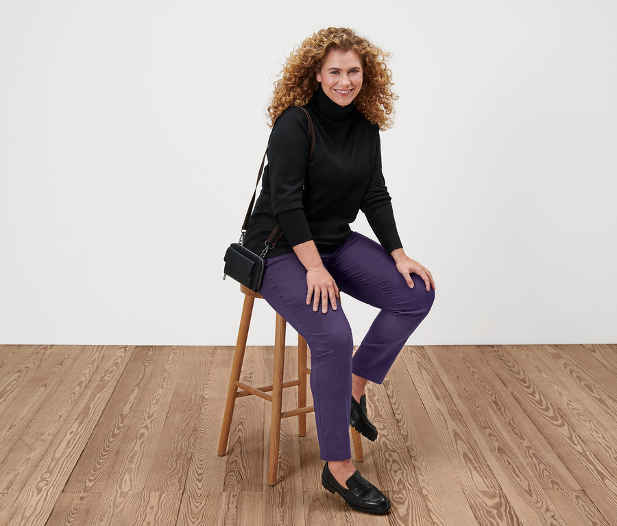 Une femme est assise sur un tabouret et porte un pull noir à col roulé en maille fine, un pantalon violet et des chaussures noires.