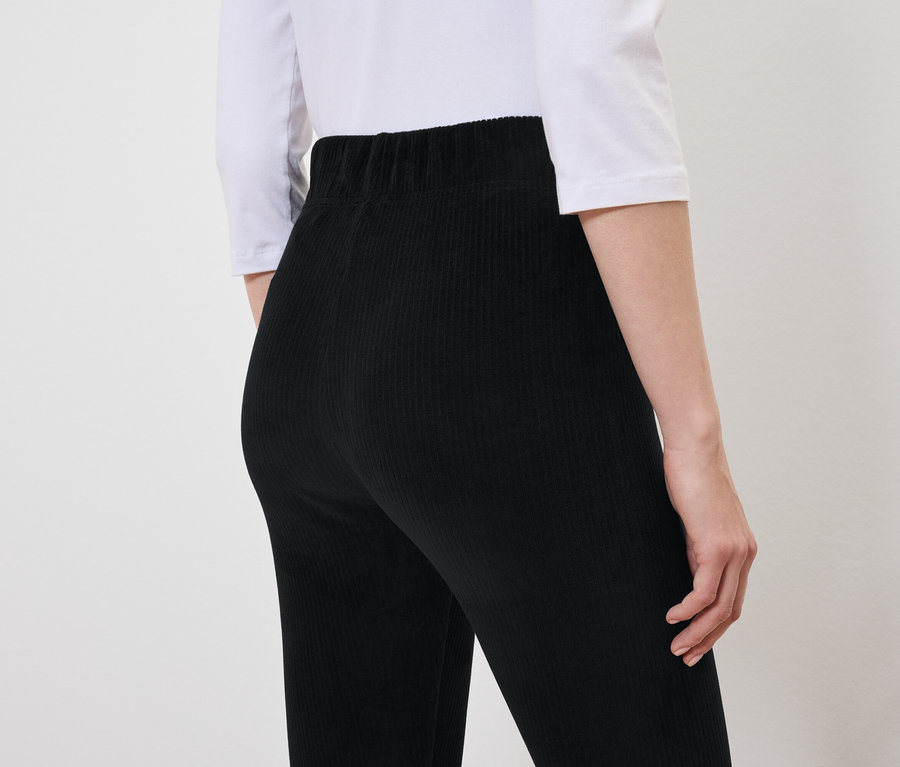 De dos, une femme porte un pantalon de velours noir et un t-shirt blanc.