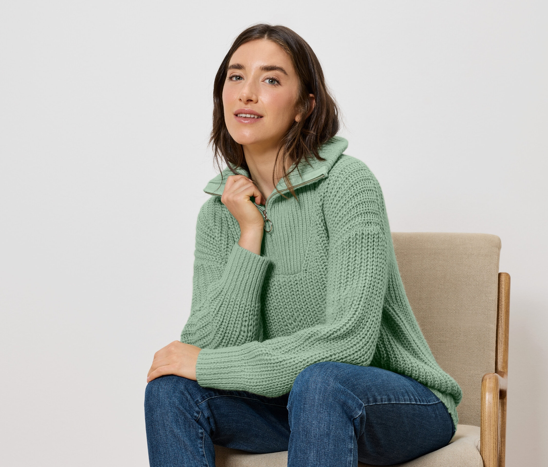 Une femme est assise sur une chaise et porte un pull vert et un jean bleu.