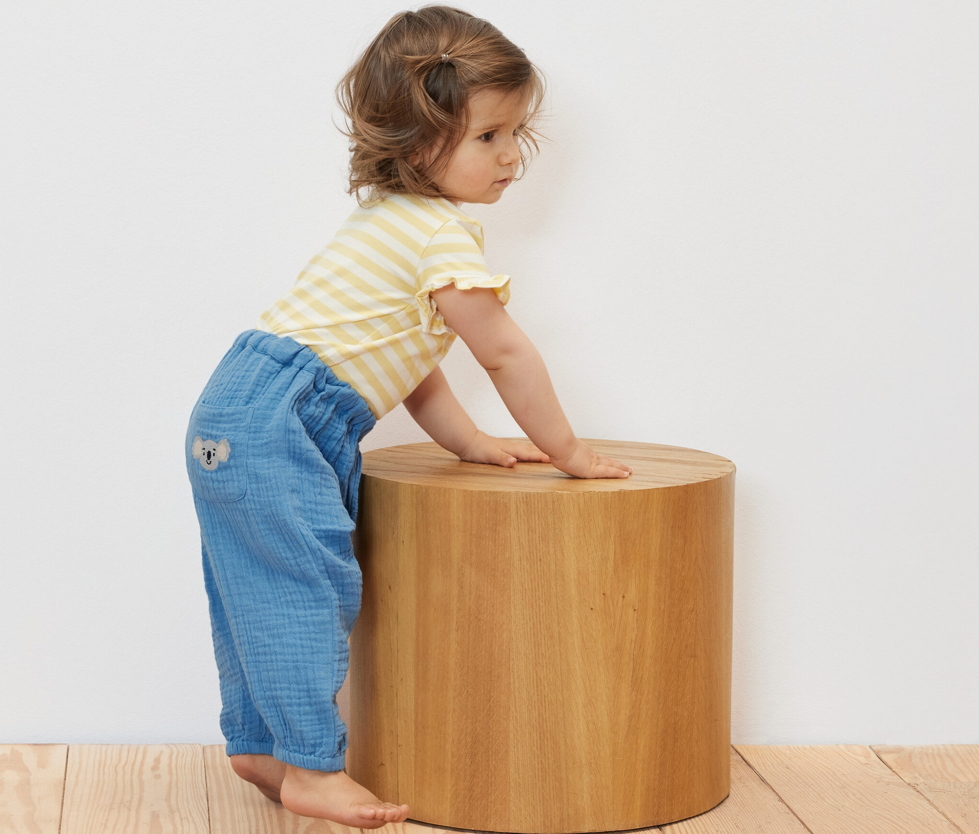 Un bébé est accoudé à un tabouret en bois et porte une culotte en mousseline pour bébé.