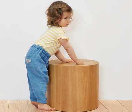 Un bébé est accoudé à un tabouret en bois et porte une culotte en mousseline pour bébé.