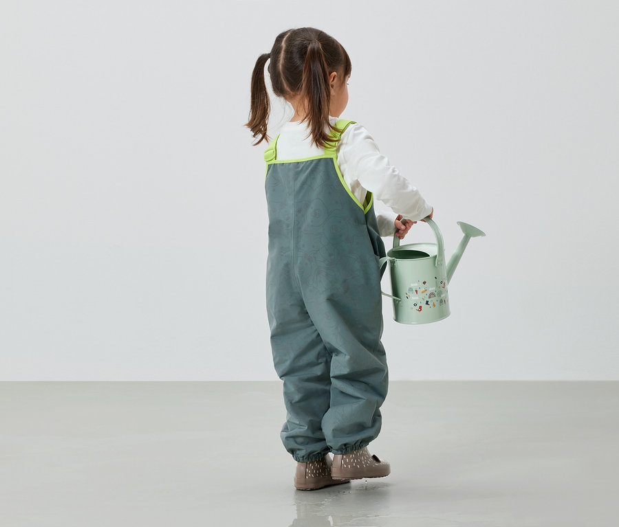 Fillette en pantalon gris tenant un arrosoir vert.