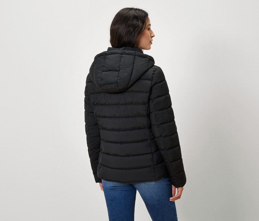 Une femme vue de dos porte une veste matelassée noire à capuche et un jean.