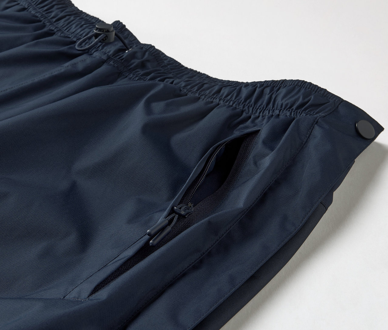Gros plan sur un pantalon de pluie high-tech avec fermeture éclair intégrale, montrant une taille élastique avec cordon de serrage et une poche zippée.