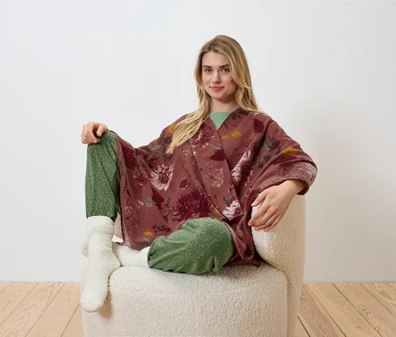 Une femme blonde est assise dans un fauteuil et porte un kimono à fleurs, un pantalon vert et des chaussettes blanches.