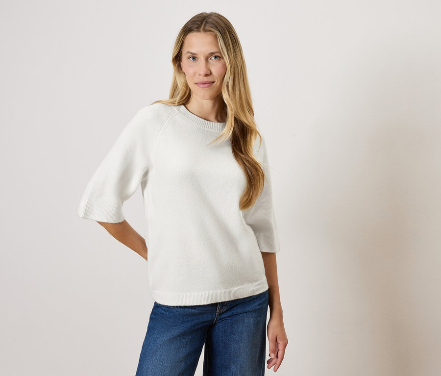 Une femme aux longs cheveux blonds pose dans un pull blanc à manches trois-quarts et un jean bleu.
