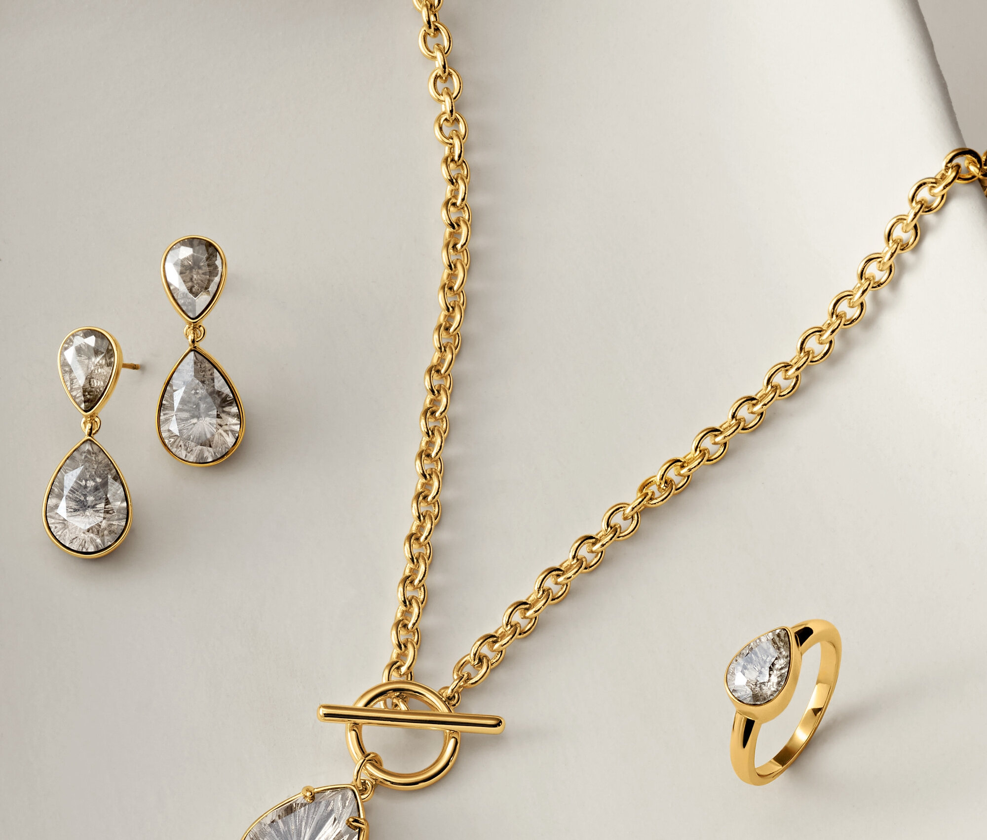 Collier, boucles d'oreilles et bague dorés avec des pierres claires en forme de larme sur fond blanc.