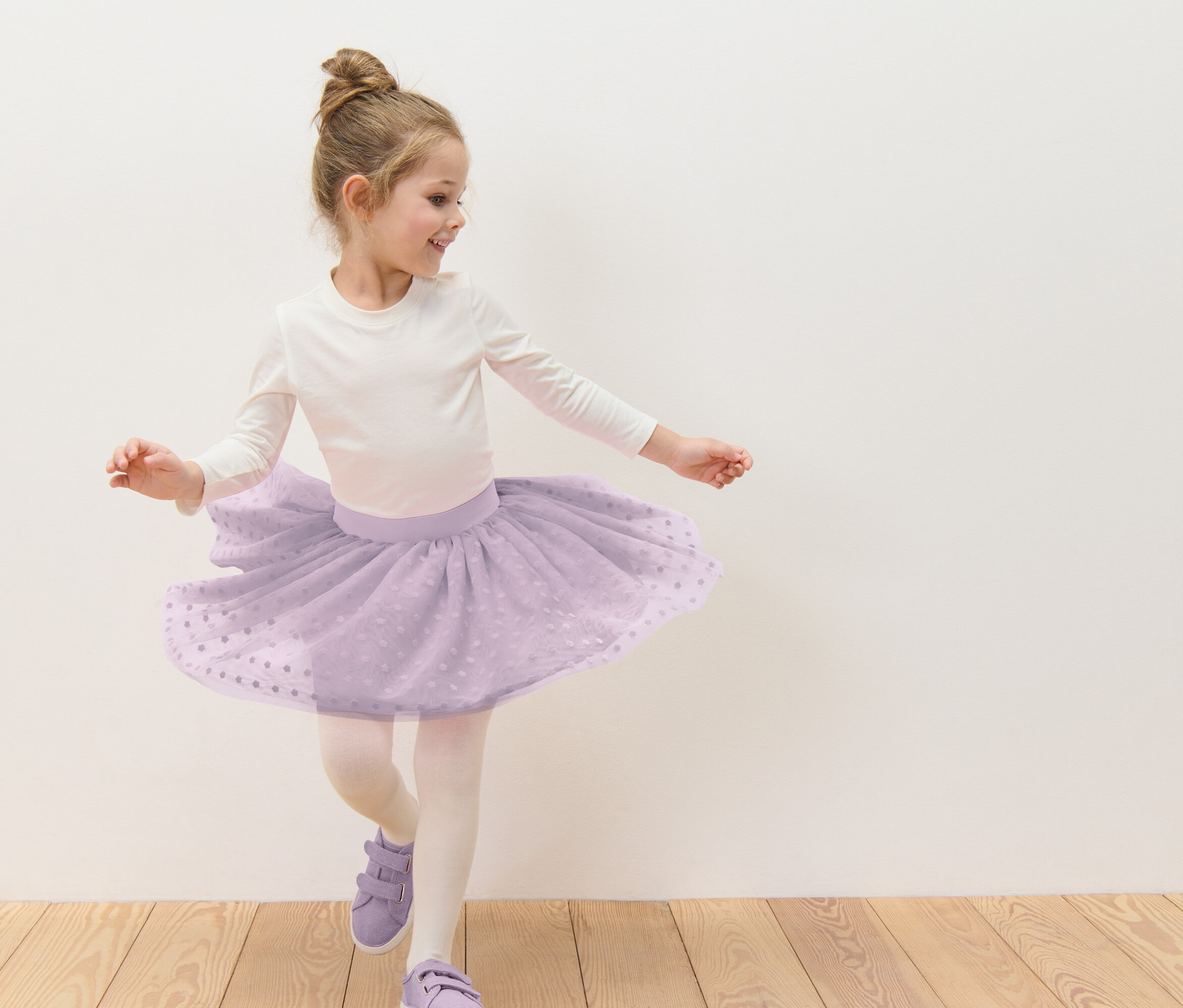 Une fillette avec un chignon danse dans une jupe en tulle violette avec des pois blancs, un t-shirt blanc à manches longues, des collants et des chaussures violettes devant un mur blanc.