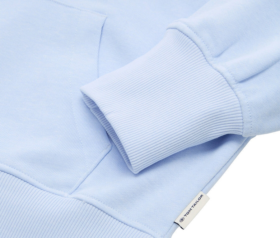 Gros plan sur un sweat à capuche bleu clair avec un bord de manche côtelé. Une étiquette Tom Tailor est visible.