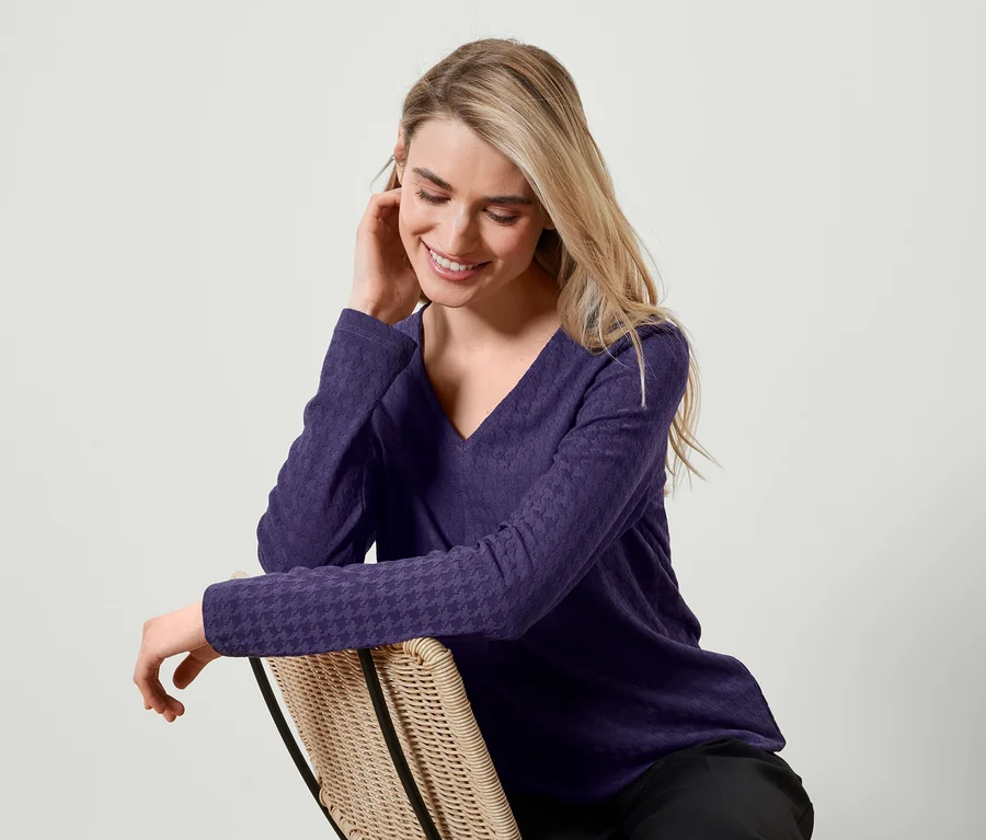 Une femme blonde est assise sur une chaise en rotin, porte un t-shirt violet à manches longues et sourit.
