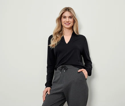 Une femme blonde portant un haut noir à manches longues et un pantalon pied-de-poule gris pose.