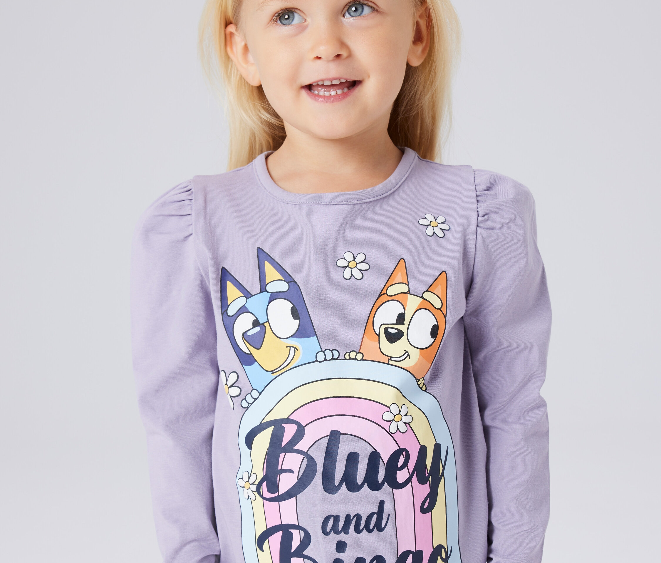 Une petite fille blonde porte un T-shirt à manches longues avec un imprimé de Bluey et Bingo sur un arc-en-ciel.