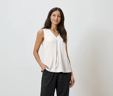 Une femme aux cheveux foncés porte un haut clair et un pantalon foncé, avec une main dans sa poche.