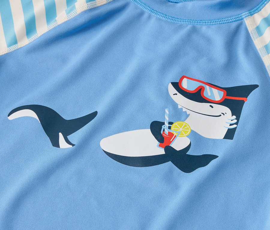Sur un tissu bleu clair, un requin portant des lunettes de soleil est représenté en train de siroter une boisson avec du citron.