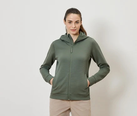 Femme portant un sweat à capuche vert olive zippé et un pantalon beige, debout avec les deux mains dans les poches.