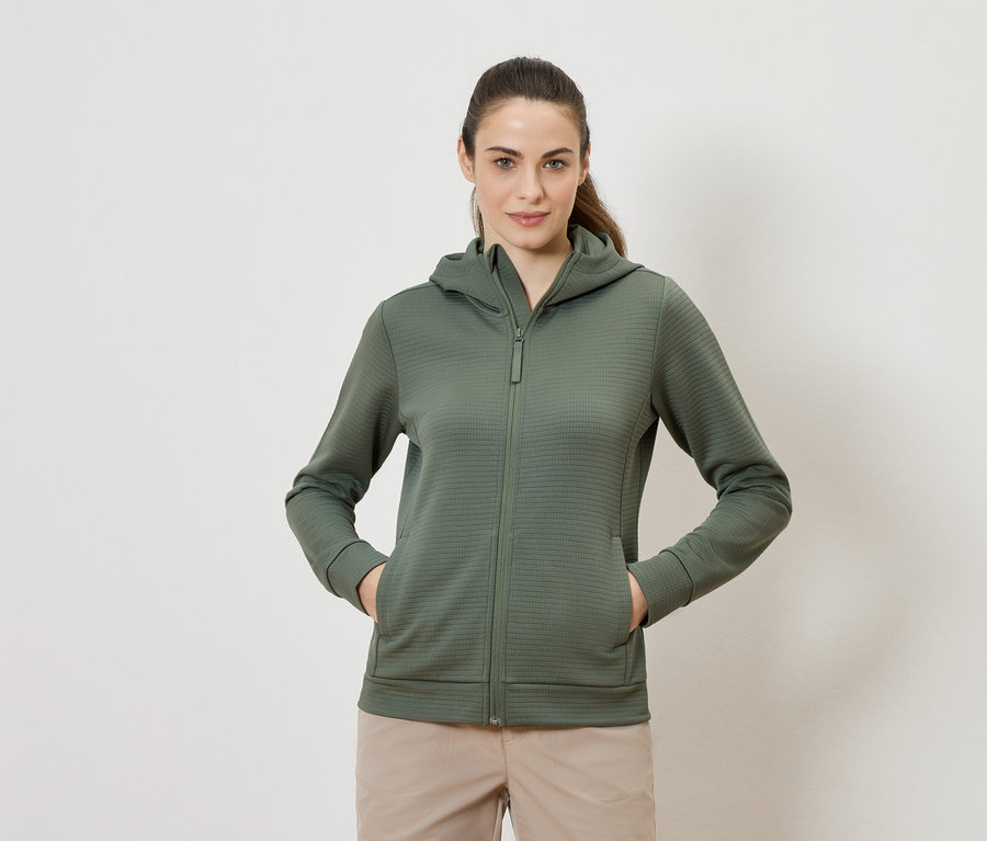 Femme portant un sweat à capuche vert olive zippé et un pantalon beige, debout avec les deux mains dans les poches.