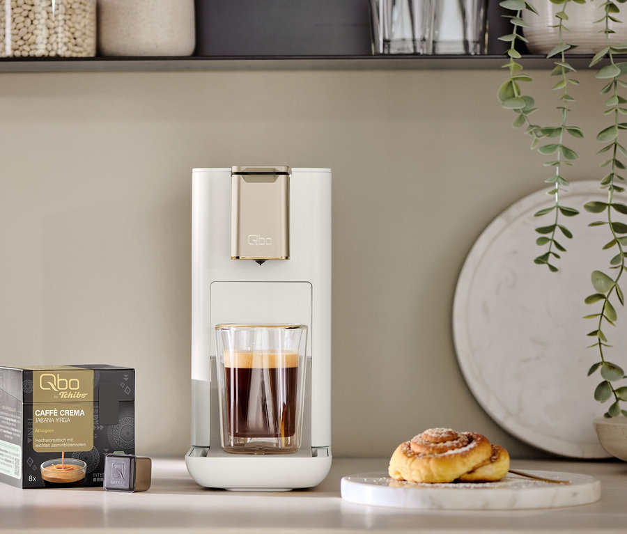 Une machine à café Qbo ICONIC Light Gold avec une tasse de café à côté. Un paquet de capsules Qbo et une viennoiserie à la cannelle sont également visibles.