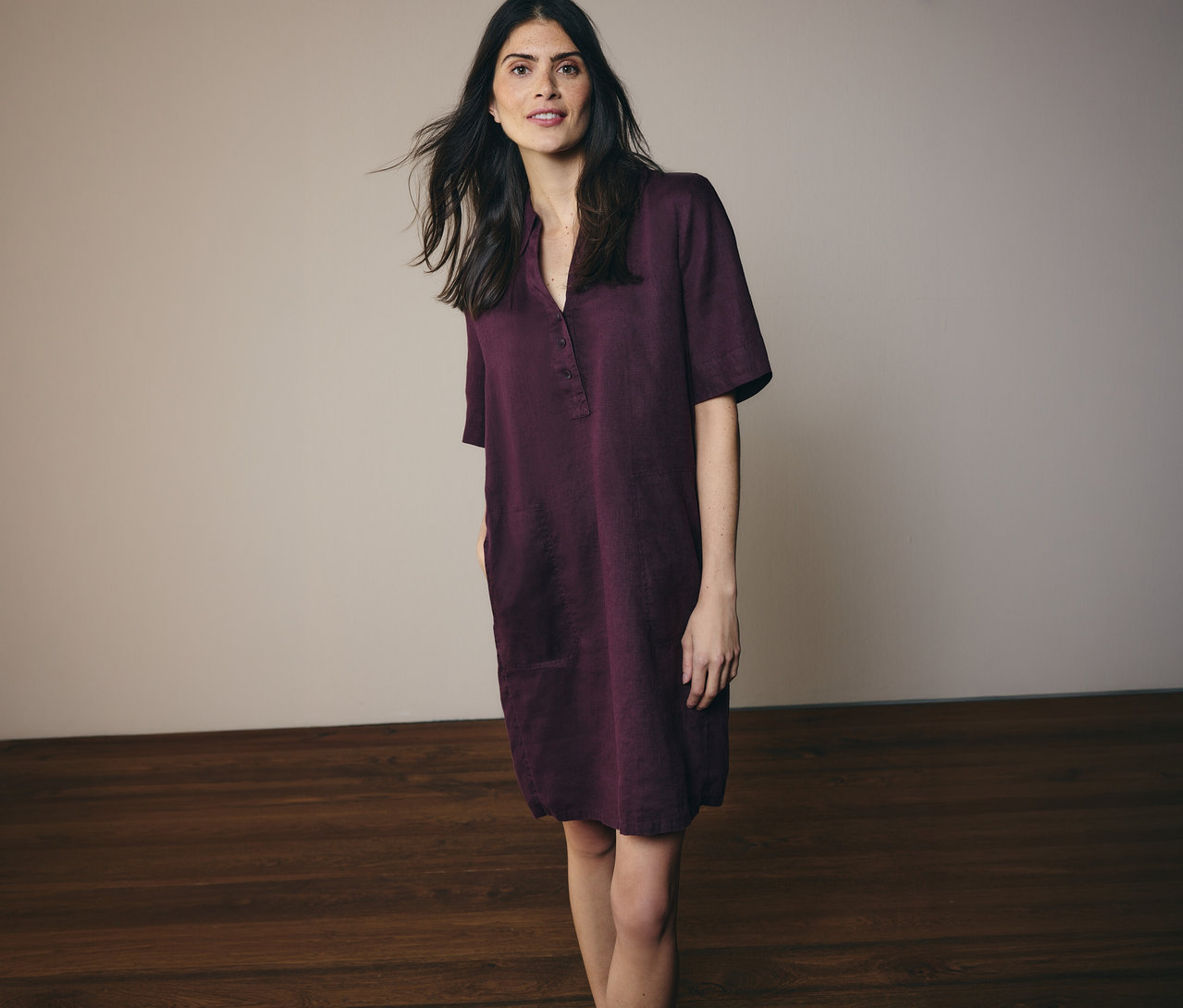 Une femme aux longs cheveux foncés porte une robe-chemise bordeaux arrivant aux genoux, avec des boutons et des poches.
