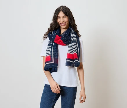 Une femme souriante aux cheveux ondulés et foncés porte un t-shirt blanc, un jean et un foulard à motifs rouge, blanc et bleu marine.