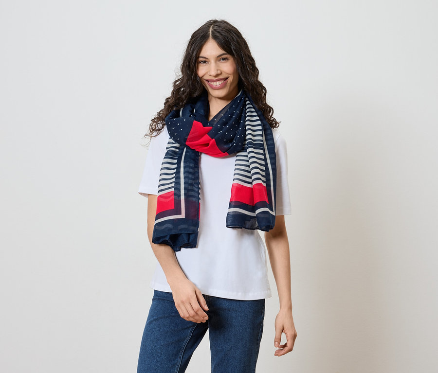 Une femme souriante aux cheveux ondulés et foncés porte un t-shirt blanc, un jean et un foulard à motifs rouge, blanc et bleu marine.