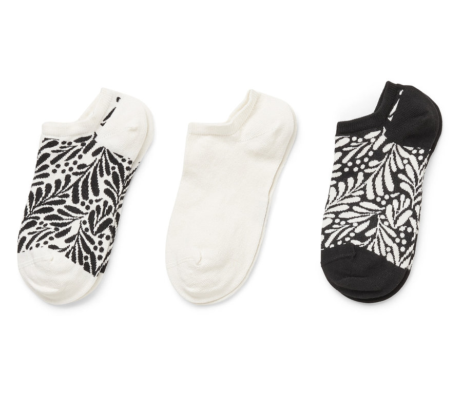 Trois chaussettes sont côte à côte : une chaussette blanche avec un motif de feuilles noires, une chaussette blanche et une chaussette noire avec un motif de feuilles blanches.