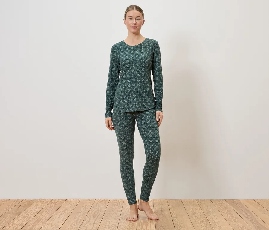 Une femme se tient pieds nus sur un plancher en bois devant un mur blanc et porte un pyjama vert foncé avec un motif vert clair.