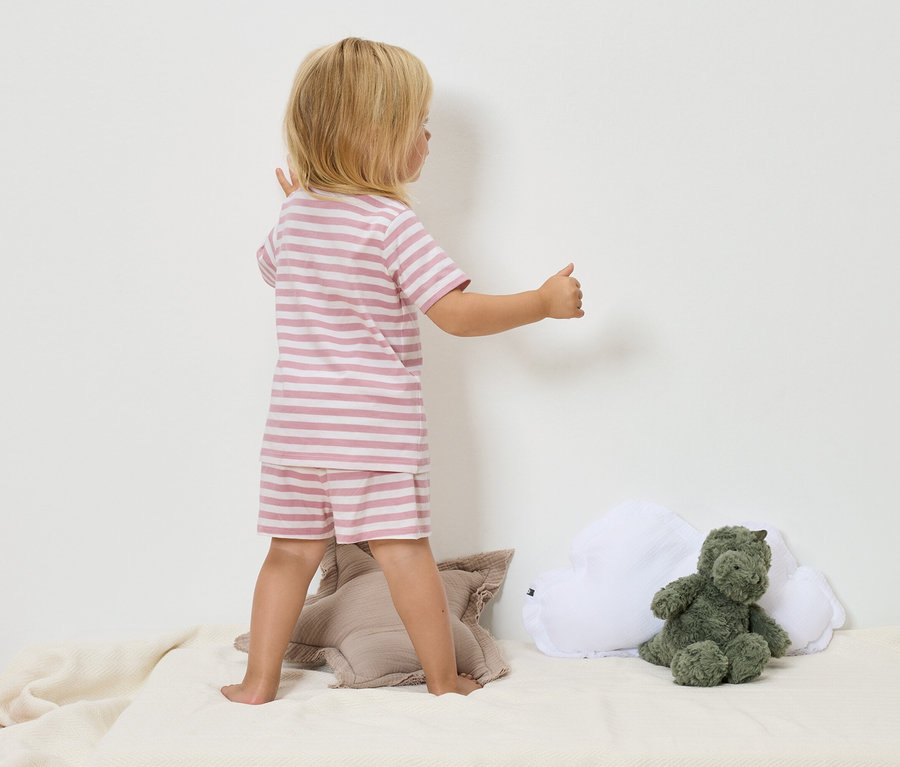 Un enfant blond portant un pyjama rayé rose et blanc se tient sur une couverture beige à côté d'une peluche de dinosaure verte et d'un coussin nuage blanc.