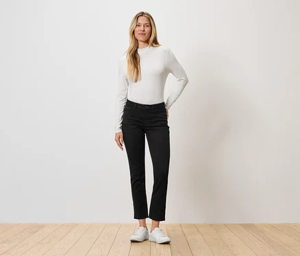 Une femme se tient devant un mur, portant un haut à manches longues, un jean noir et des baskets blanches.