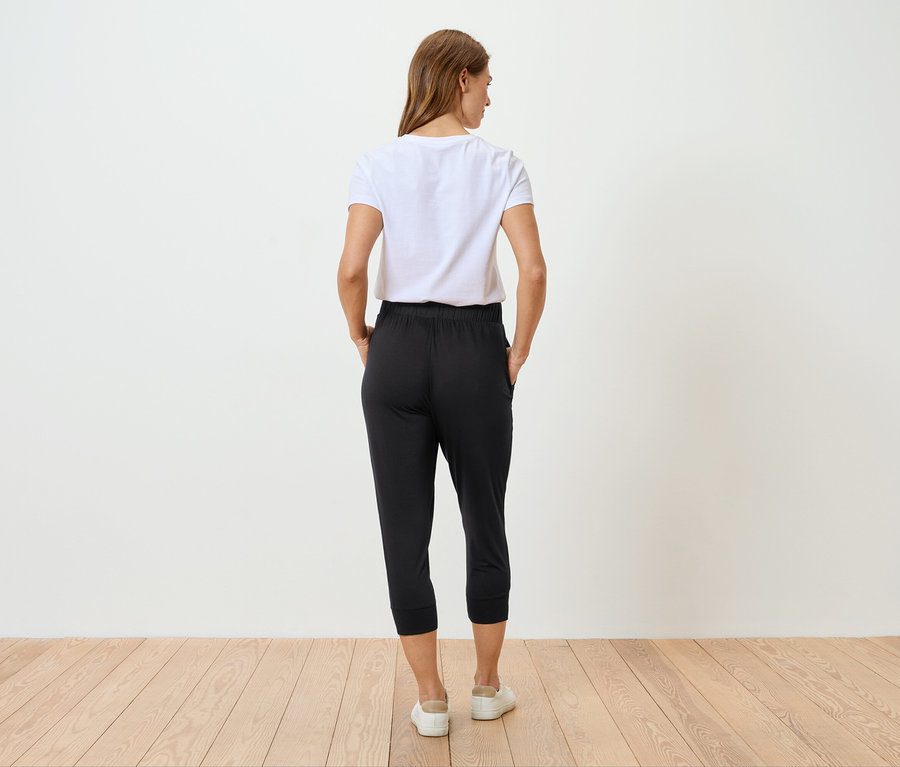 Femme vue de dos portant un t-shirt blanc et un pantalon noir, debout sur un plancher en bois.