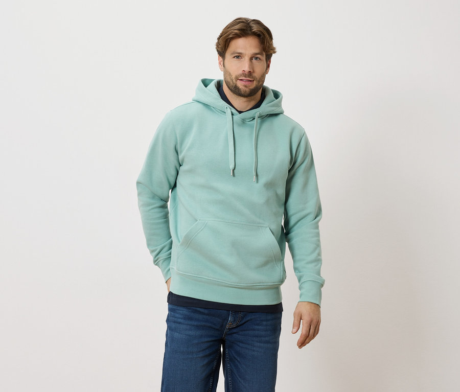 Homme portant un sweat à capuche turquoise clair et un jean debout devant un fond blanc.