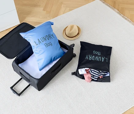 Sur un tapis se trouvent une valise ouverte avec un sac à linge bleu à l'intérieur, à côté d'un sac à linge noir et d'un chapeau.