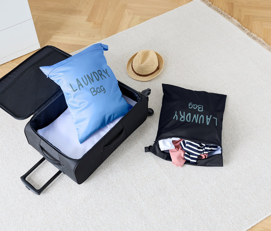 Sur un tapis se trouvent une valise ouverte avec un sac à linge bleu à l'intérieur, à côté d'un sac à linge noir et d'un chapeau.