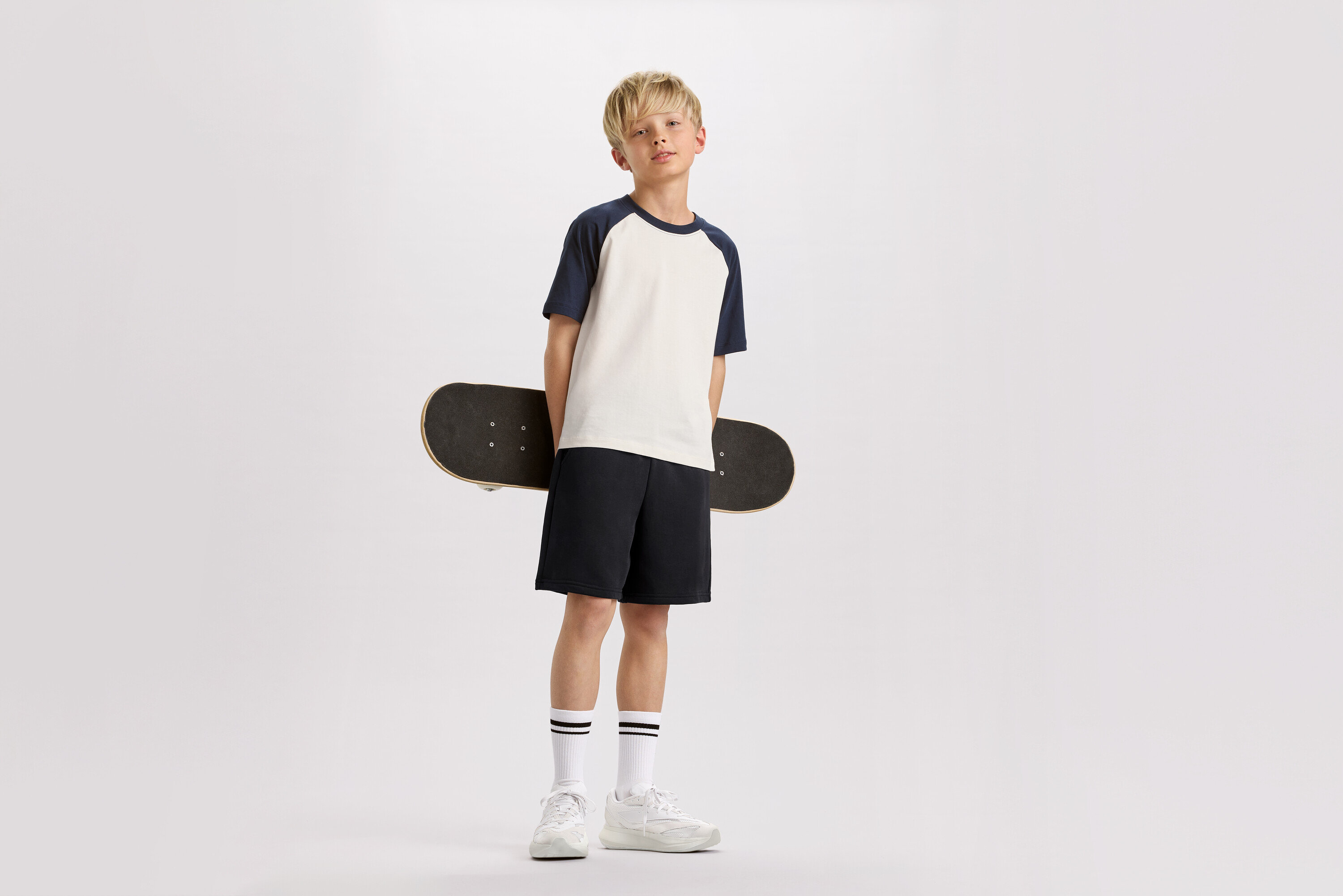 Un garçon blond tient un skateboard dans son dos, vêtu d'un t-shirt à manches bleu marine, d'un short et de baskets.