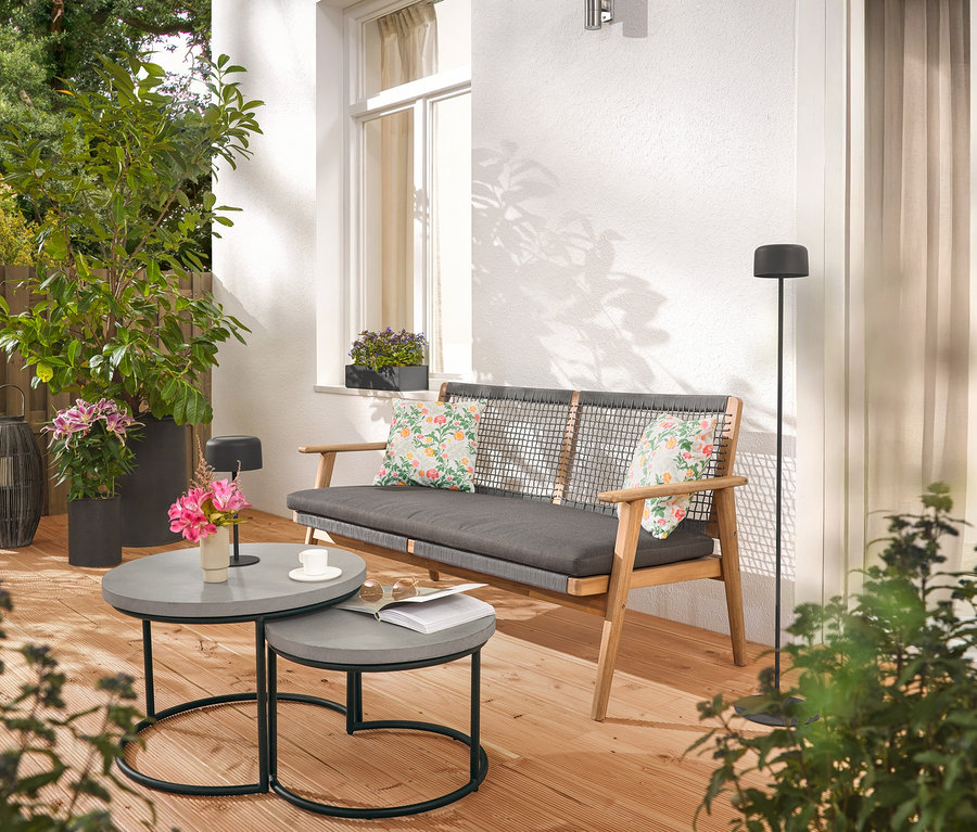 Sur une terrasse, un canapé avec des coussins, deux tables grises, un lampadaire et des plantes.