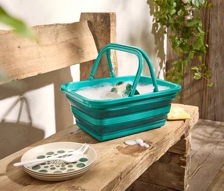 Un seau pliable vert rempli d'eau mousseuse est posé sur un banc en bois, à côté d'assiettes et de couverts.