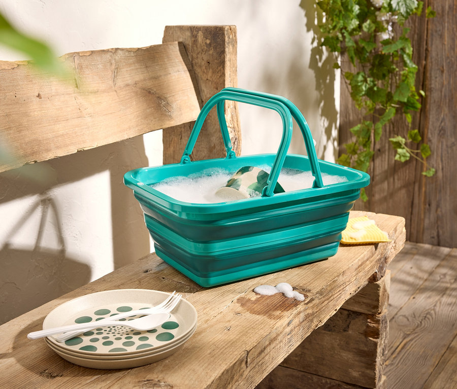 Un seau pliable vert rempli d'eau mousseuse est posé sur un banc en bois, à côté d'assiettes et de couverts.
